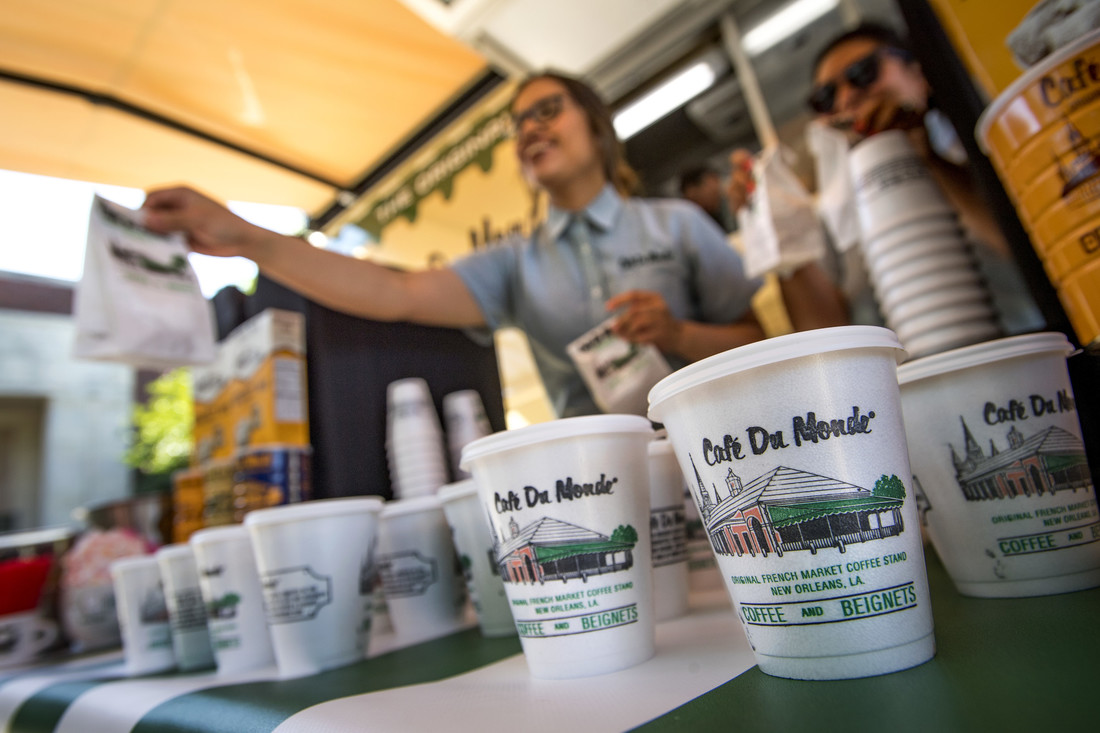 Cafe Du Monde coffee cups lined up on counter with smiling worker passing a white bag to customer.